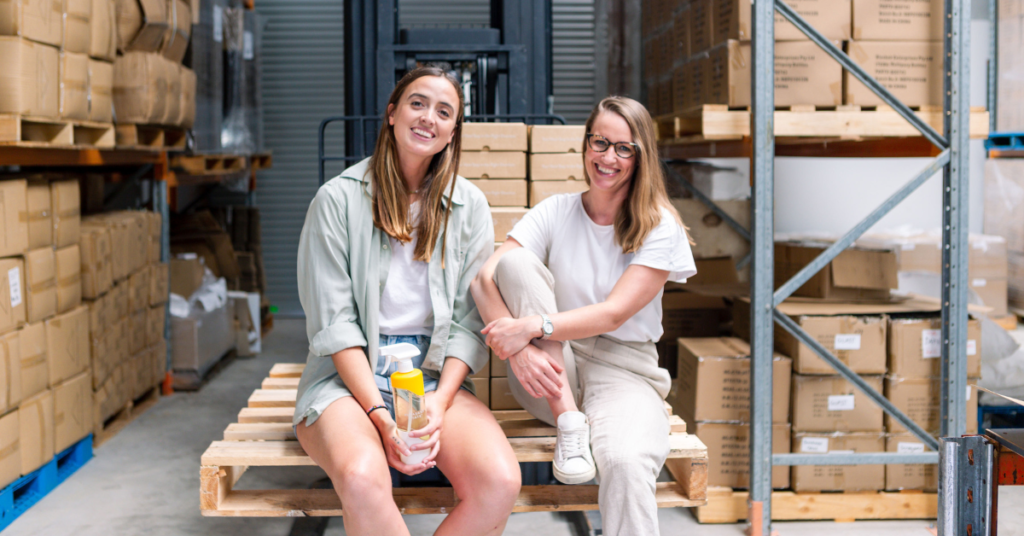 Two people smiling and looking directly at the camera while sitting on a pallet in a warehouse