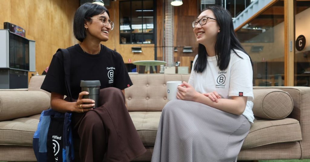 Two girls talking and drinking coffee. Both are wearing t-shirts with the certified B Corporation logo.