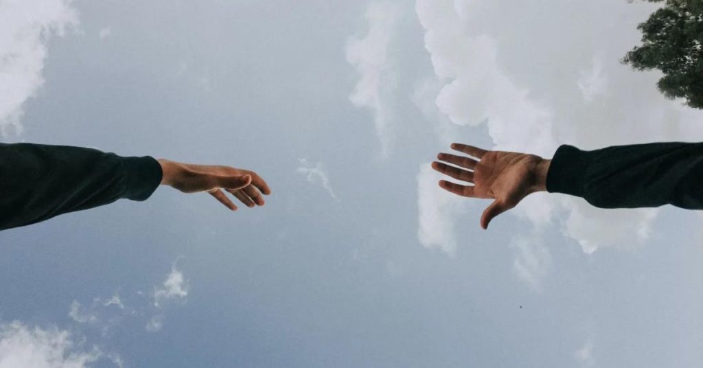 Two hands reaching towards each other in front of the sky