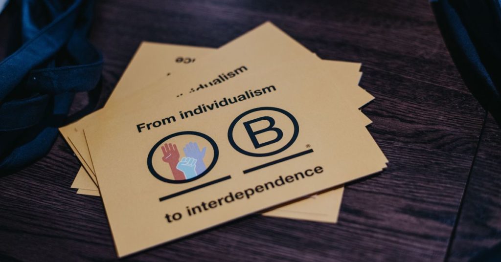 A yellow B Corp postcard which reads 'from individualism to interdependence' with the B Corp logo and three hands