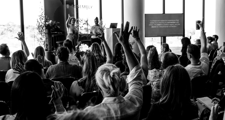 Black and white image of group of people raising their hands facing towards a platform with two speakers interacting with the attendees of the B Corps Assembly session 2024