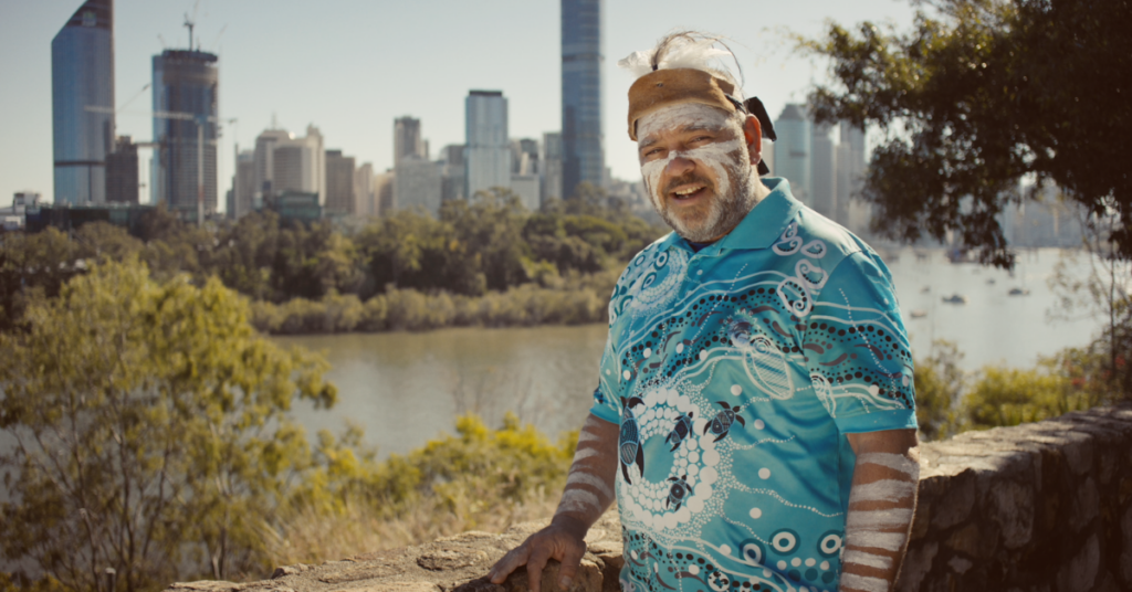 New B Corp Carbon creative work with AusPost to show a picture of a First Nations man wearing a blue coloured t-shirt with Aboriginal art print on it. He has white chalk paint on his face and Melbourne city view is in the background