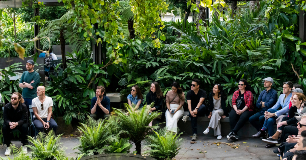 New B Corp Hassell team sitting in an outside circular seated place with lots of green plants and trees around them