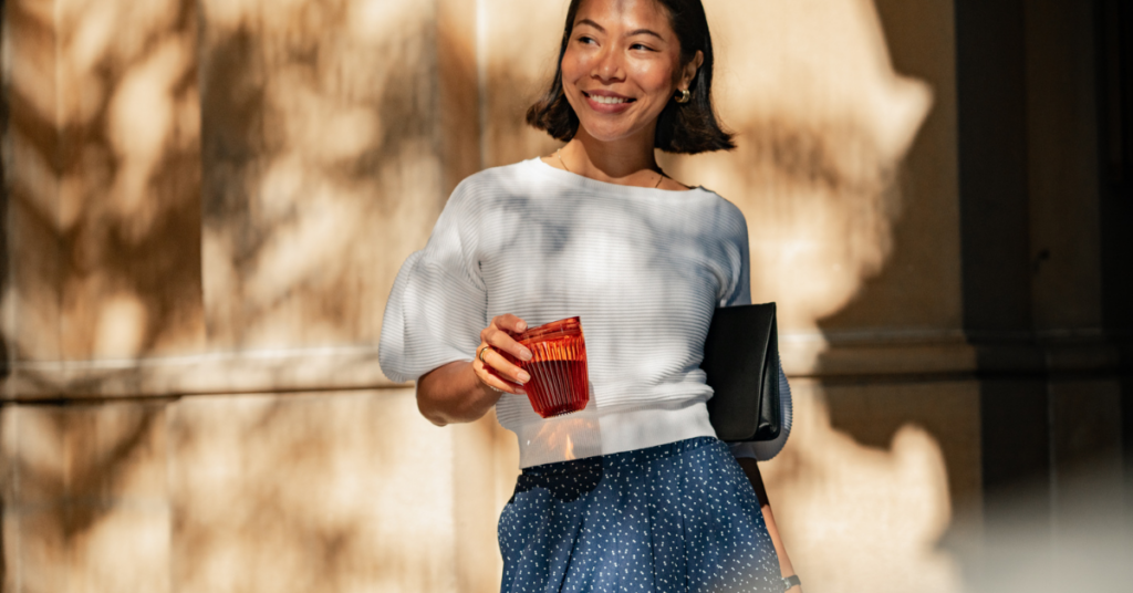 A woman is holding a red colour Certified B Corporation Huskee cup in her right hand. The woman is wearing a white blouse and a blue skirt, carrying a black cluck bag in her left arm. The background is a beige coloured wall with leaves shadow falling on it.