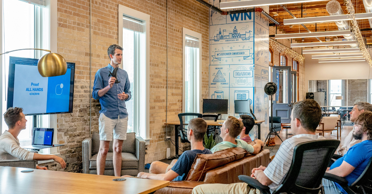 A group of people in a meeting room with a Man in the front wearing a blue shirt and grey pair of shorts. He is holding a leadership award in his hand and addressing the people sitting in the room. There are seven people in the room. Three are sitting on a couch and the rest are sitting on chairs. There's a screen behind the standing man which says " Proof, All Hands". 