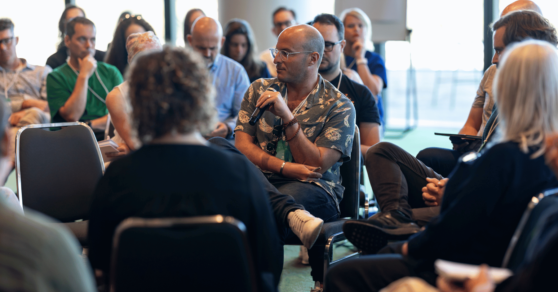 A B Council member holds a microphone while speaking during an interactive session. Other B Council members are seated in a circle, listening closely and engaged in the discussion.