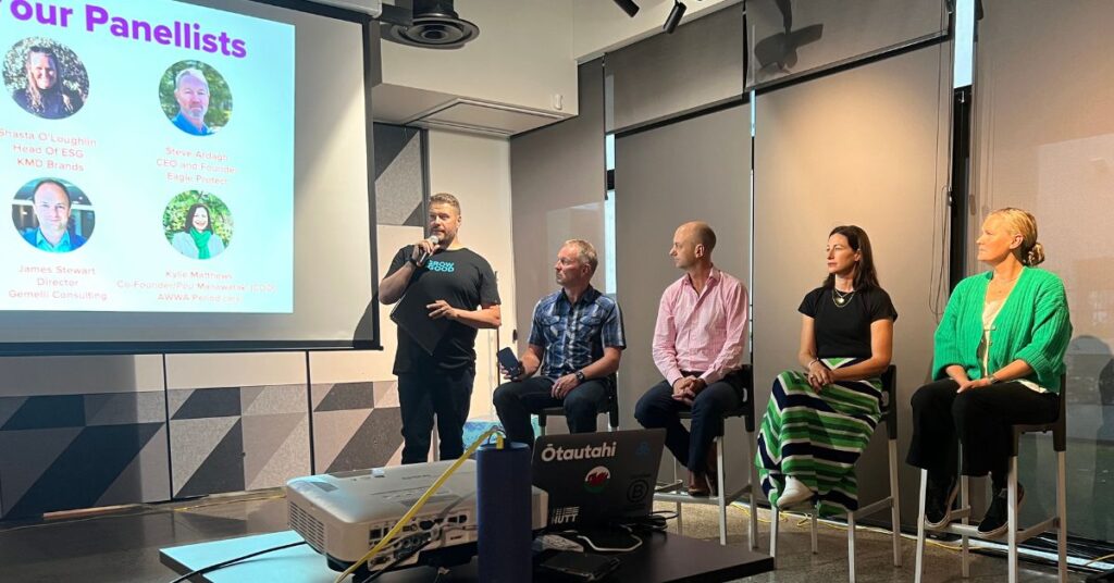 One man standing with a microphone next to a presentation, four other panelists sitting