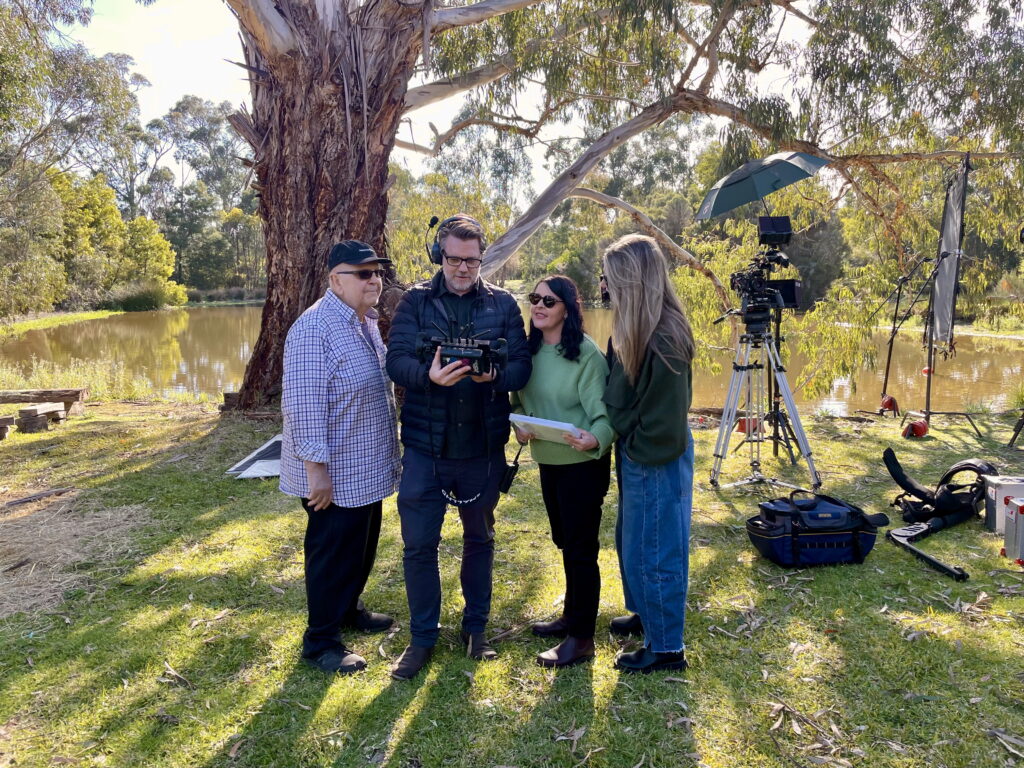 4 people looking at a camera. The film crew are set up in a park with a lake behind.