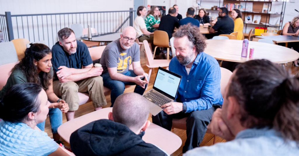 Seven people sat in a circle looking at one of the people’s laptop screens.