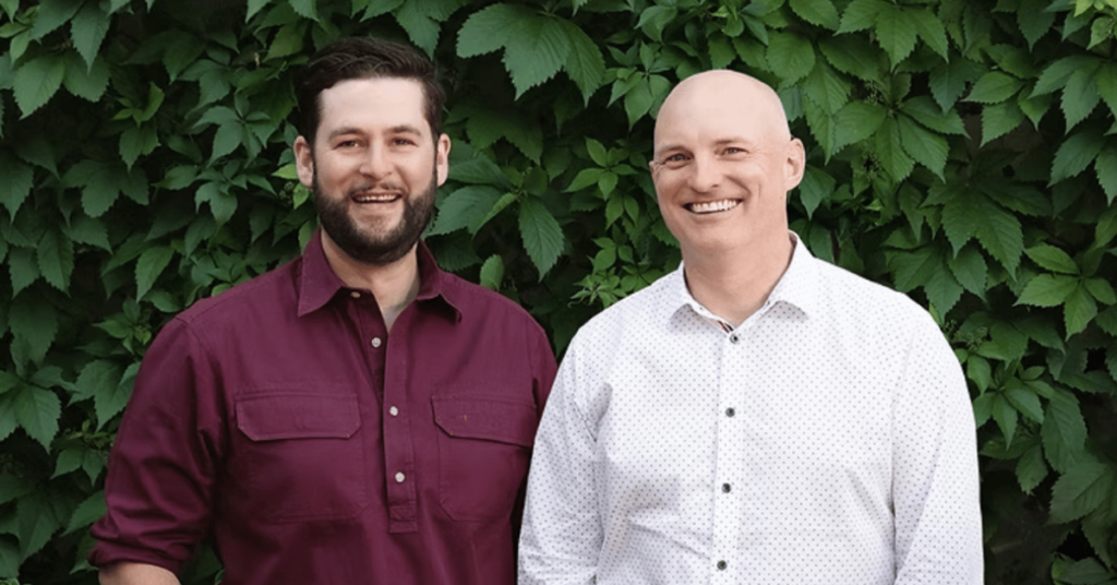 Two people stood smiling at the camera, in front of a wall covered in leaves