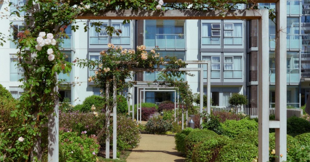 A block of flats at the end of a pathway, which has arches and rose vines across it.