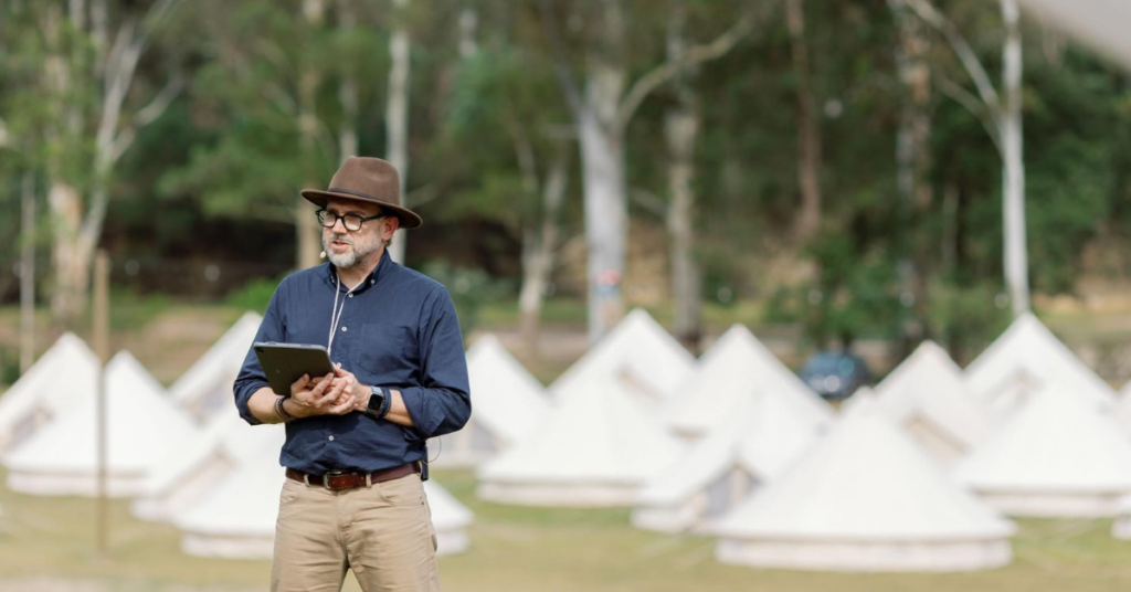 Speaker standing outdoors with a tablet and headset microphone, with rows of tents in the background.