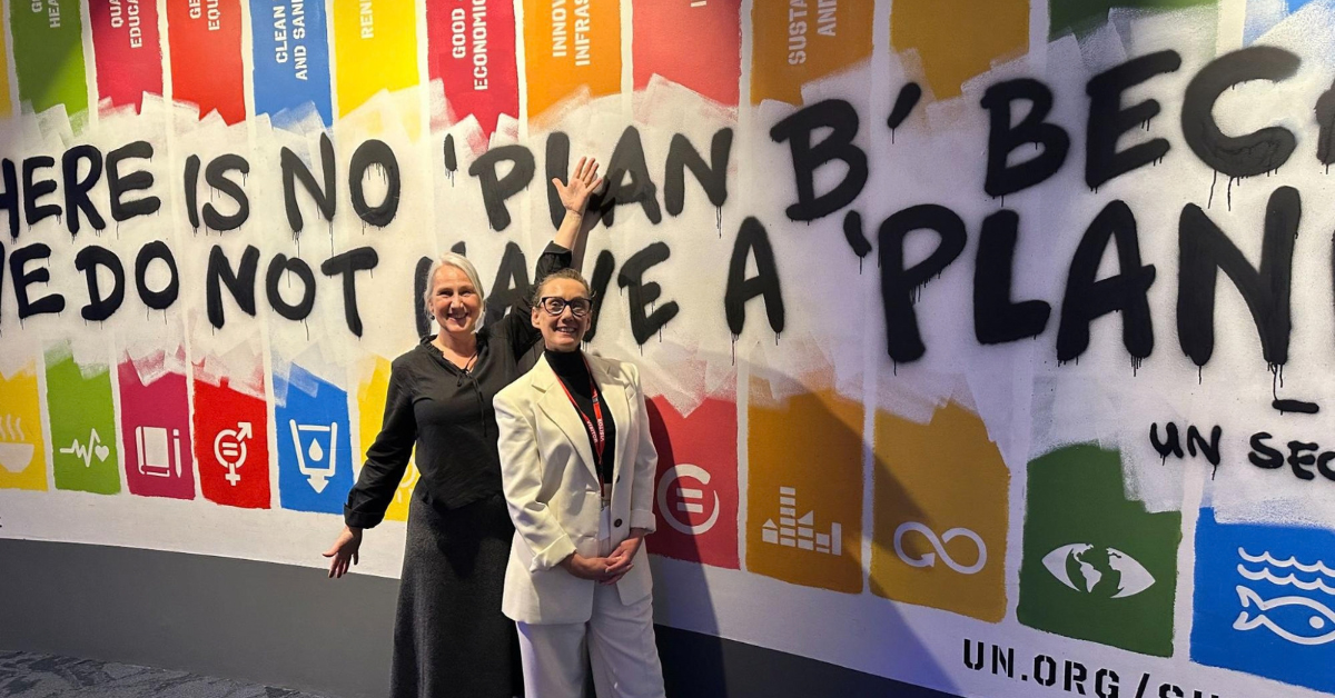 Andrea Spencer-Cooke and one other person stood in front of a picture of the UN sustainability goals.