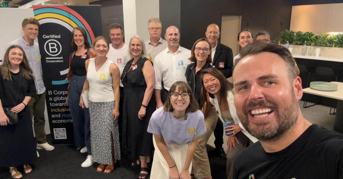Andrea Spencer-Cooke and Lauren Diggle with members of the B Corp community stood in front of a Certified B Corp logo on a banner.