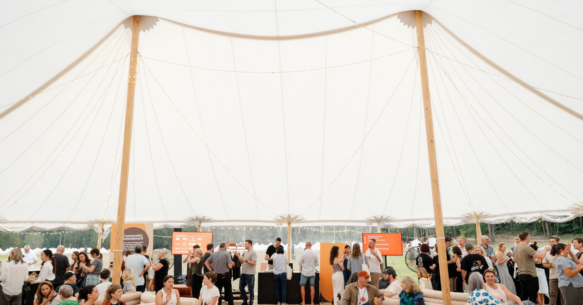 A group of people sit on couches and stand under a giant airy white tent, at a conference.