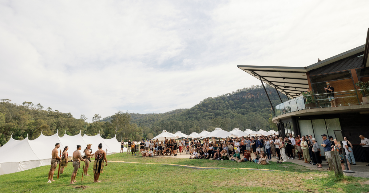 A group of people stands outside in a vast landscape while a group of five performs a haka.