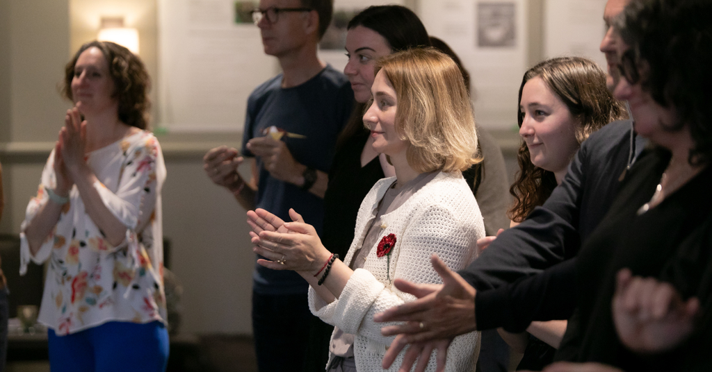 A group of people stand watching a speaker, clapping. 