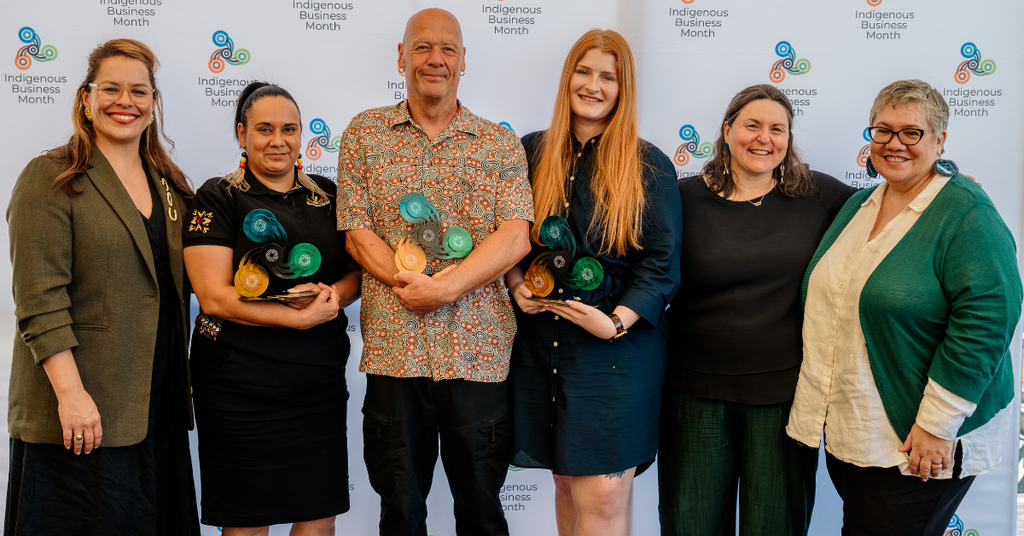 A group of six people stand in a line, with three holding awards in the style of the Indigenous Business Month logo.