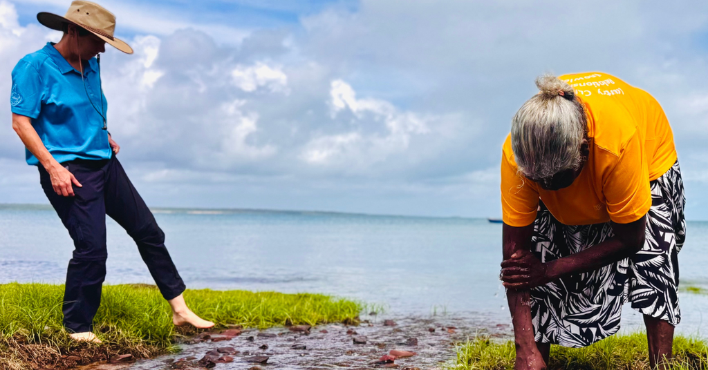 One person stands barefoot on the edge of a river inlet, and another leans over into the grass. 