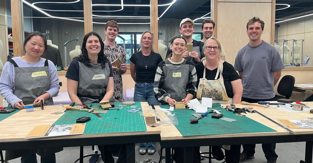 A group of 9 people smile while sitting at a craft table, wearing aprons and working on textiles.