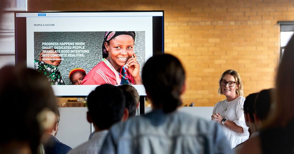 A woman presents a People & Culture presentation that reads: Progress happens when smart, dedicated people translate good intentions into concrete realities. 